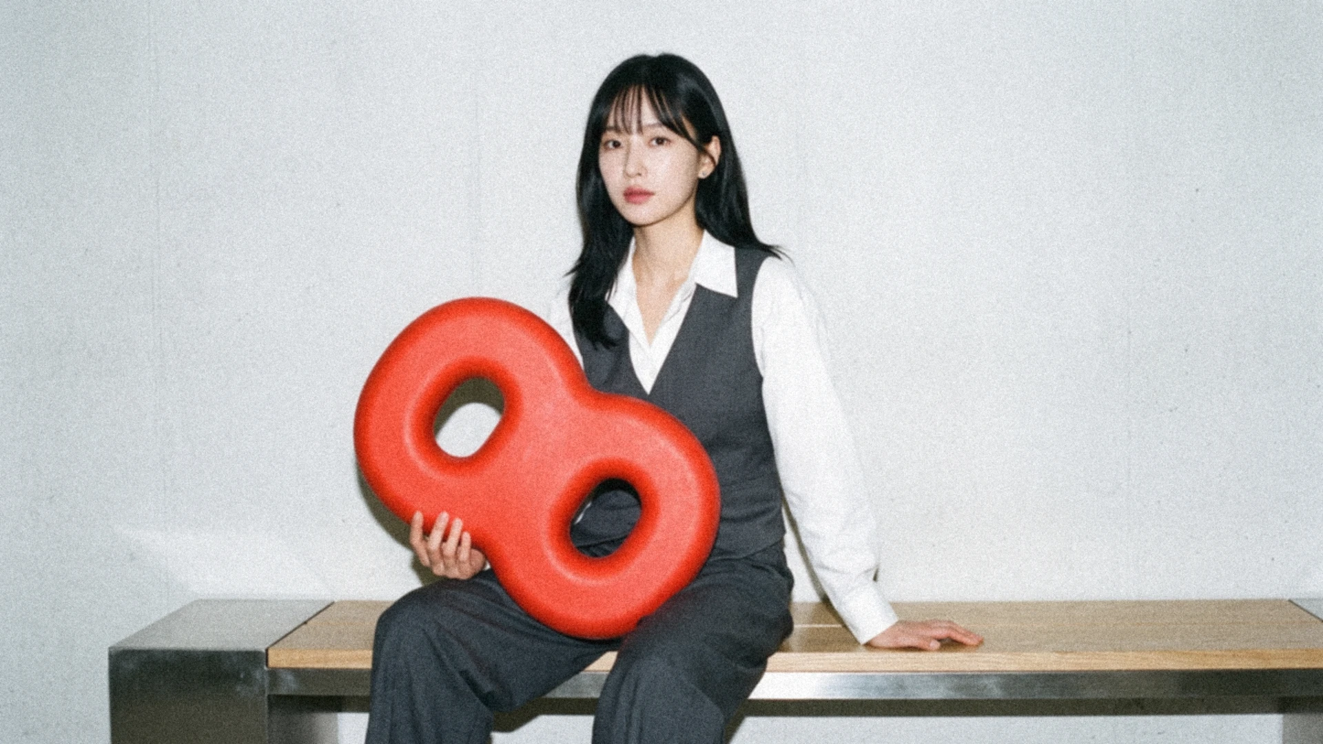 A woman in a white blouse and gray waistcoat sits on a bench, holding a vermilion double-loop Link shape.