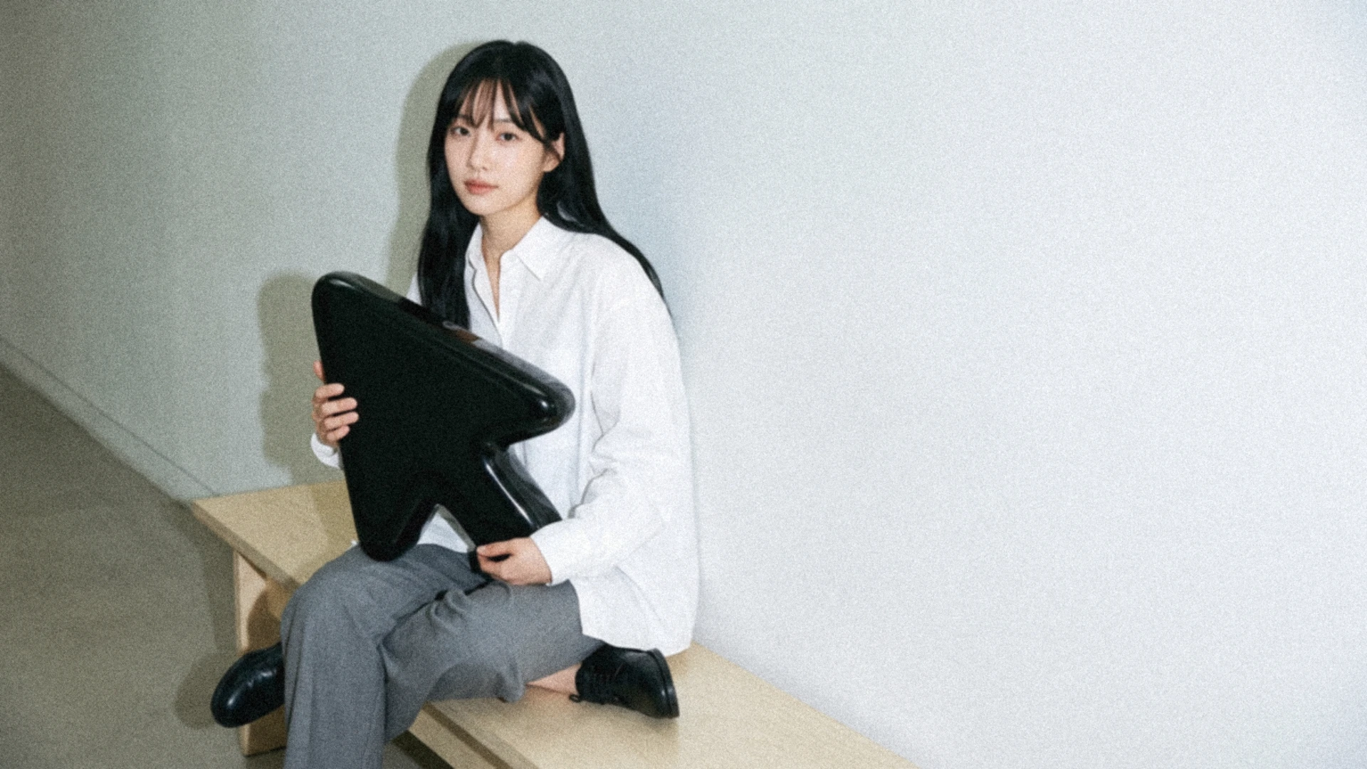 A young woman with long black hair and a white shirt sits on a pale wooden bench in a bright corridor, holding an oversized glossy black cursor on her lap.