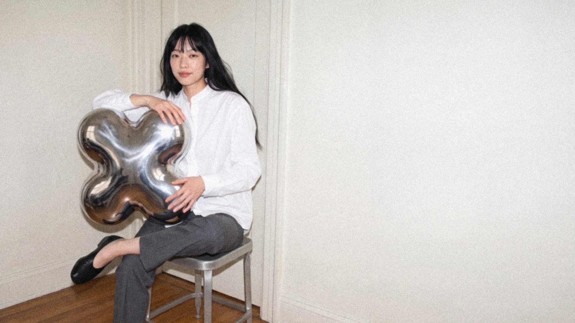 A woman in a white blouse sits on a metal stool in a doorway, holding an oversized four-lobed chrome Plump shape.