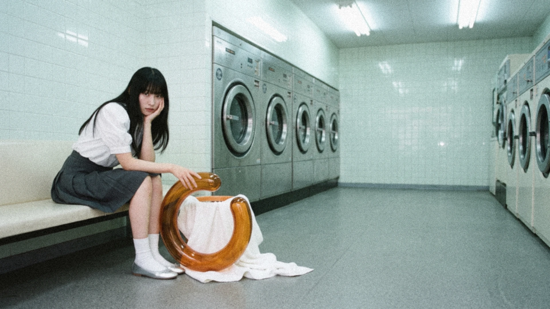 A woman with long black hair waits in a cool green laundromat beside an amber glass Time ring, with soft focus and film grain.